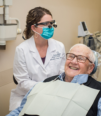 Female dentist with mask and magnification glasses sitting next to male patient