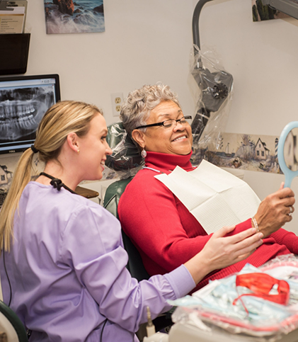 Older female patient checking smile in mirror with dental team member seated nearby