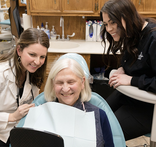 Senior woman in dentla chair checking smile in mirror while being watched by dental team